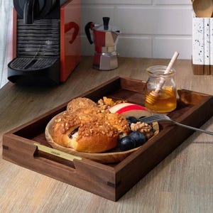 May include: A wooden serving tray with a plate of food, including a pastry, fruit, and nuts. A small jar of honey with a honey dipper sits next to the tray. A coffee machine and a stovetop espresso maker are in the background.