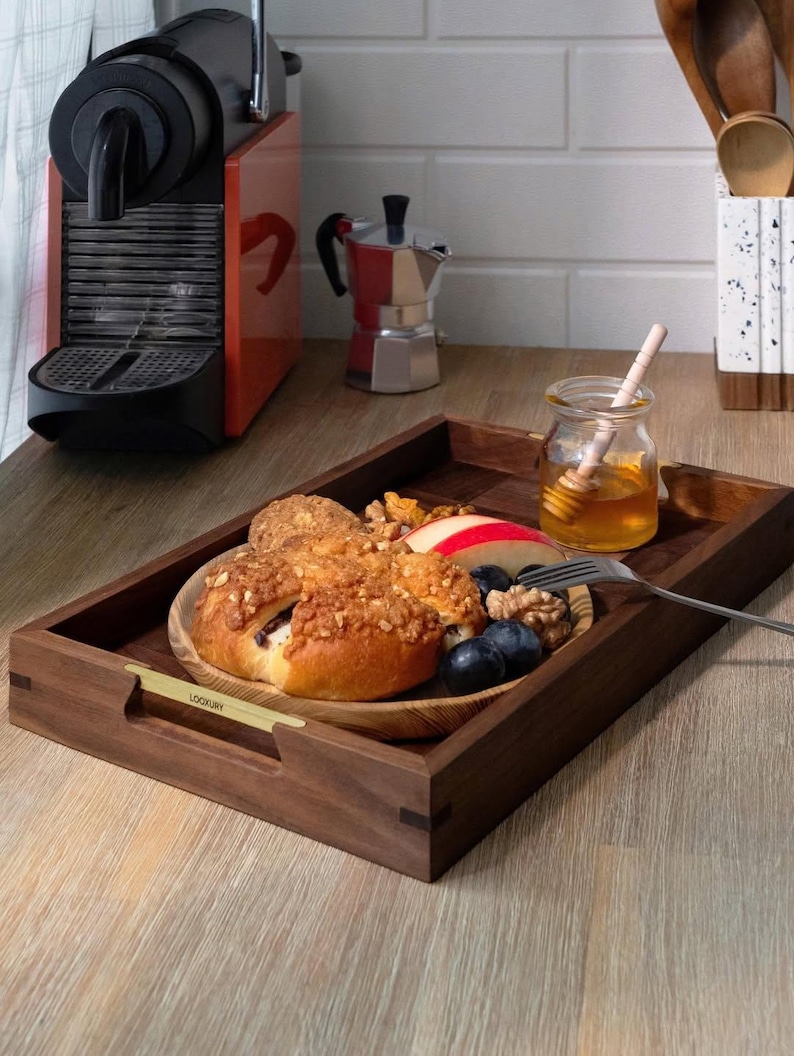 May include: A wooden serving tray with a plate of food, including a pastry, fruit, and nuts. A small jar of honey with a honey dipper sits next to the tray. A coffee machine and a stovetop espresso maker are in the background.