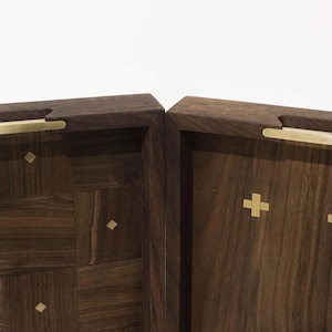 May include: Two wooden boxes with a dark brown finish. One box features a geometric pattern, while the other has gold cross inlays. Both boxes have gold-colored handles.
