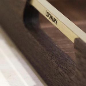 May include: Close-up of a dark brown wooden tray with a light-colored handle. The handle has the word "LOOXURY" printed on it in black. The tray has a smooth, polished finish.