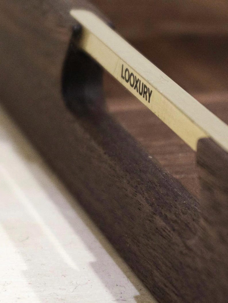 May include: Close-up of a dark brown wooden tray with a light-colored handle. The handle has the word "LOOXURY" printed on it in black. The tray has a smooth, polished finish.
