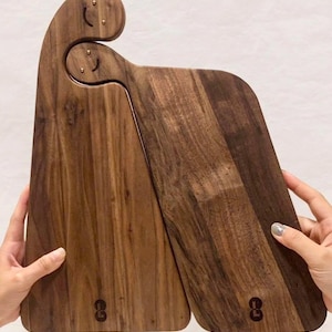 May include: Two wooden cutting boards with a unique design. The boards are made of dark brown wood and feature a curved handle with small brass accents. Each board has a rounded bottom and a logo near the base. The boards are held open by hands.