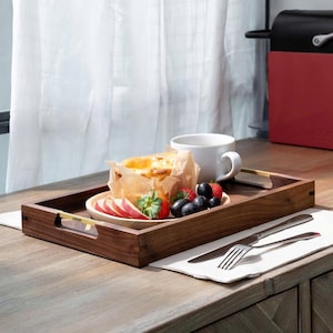 May include: A wooden serving tray with a handle, holding a plate of food, fruit, and a white mug. The tray has a dark brown finish and gold-colored handles. The plate contains a pastry, sliced apples, strawberries, and blueberries. A knife and fork rest on a napkin.