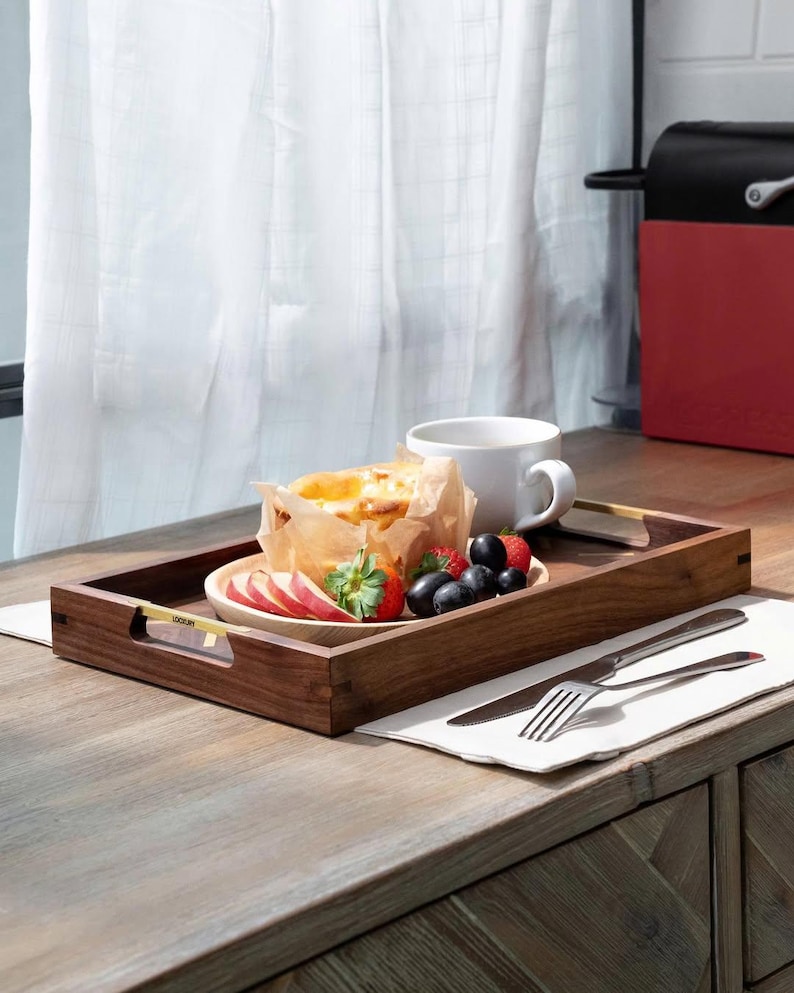 May include: A wooden serving tray with a handle, holding a plate of food, fruit, and a white mug. The tray has a dark brown finish and gold-colored handles. The plate contains a pastry, sliced apples, strawberries, and blueberries. A knife and fork rest on a napkin.
