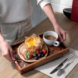 May include: A wooden serving tray with a pastry, fruit, and coffee. The tray has a geometric design and is holding a small wooden plate with sliced apples, strawberries, and blueberries. A white mug of coffee and silverware are also present.