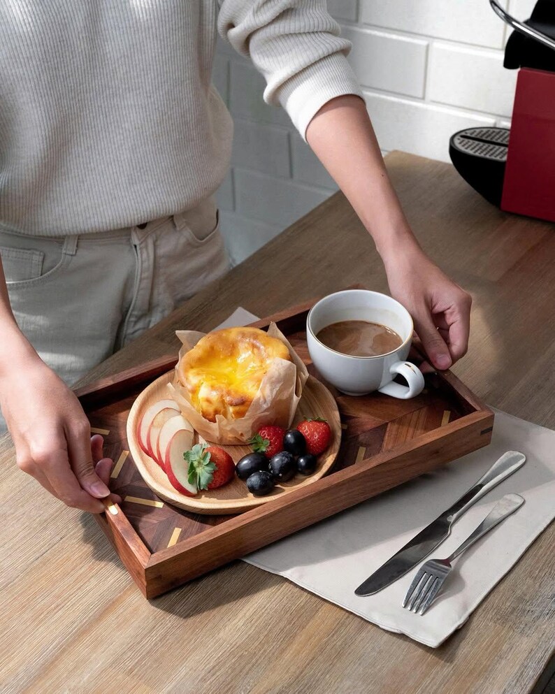 May include: A wooden serving tray with a pastry, fruit, and coffee. The tray has a geometric design and is holding a small wooden plate with sliced apples, strawberries, and blueberries. A white mug of coffee and silverware are also present.