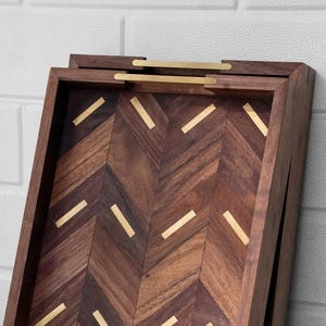 May include: A wooden serving tray with a herringbone pattern and brass accents. The tray is rectangular with a dark brown finish and gold-colored handles. The interior features a chevron design with gold inlays, adding a touch of elegance.