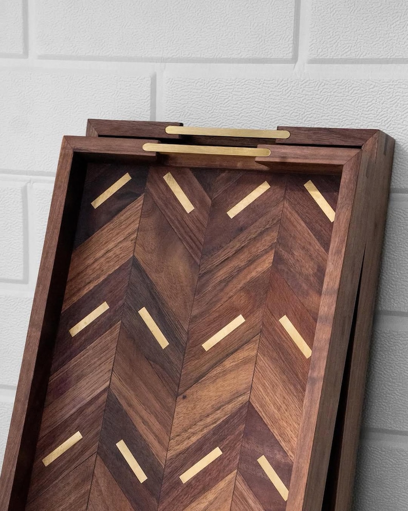 May include: A wooden serving tray with a herringbone pattern and brass accents. The tray is rectangular with a dark brown finish and gold-colored handles. The interior features a chevron design with gold inlays, adding a touch of elegance.