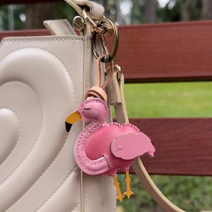 May include: A pink flamingo keychain with a yellow beak and legs, and a small pink cloud-shaped wing. The keychain is attached to a beige leather strap and a gold-colored clasp. The flamingo is stitched with a white thread.
