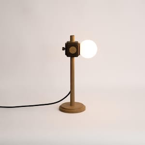 May include: A contemporary wooden desk lamp with a spherical light bulb. The lamp has a dark square head with a small knob and a wooden base. A black power cable extends from the base.