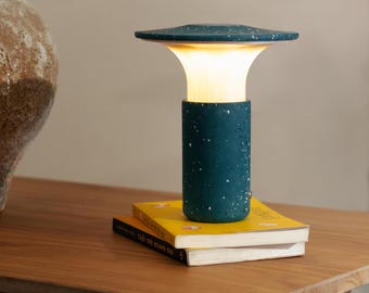 May include: A teal-colored table lamp with a speckled pattern, emitting a warm glow. The lamp has a cylindrical base and a flat, disc-shaped top. It rests on a stack of yellow books on a wooden surface. The lamp's design is modern and minimalist.