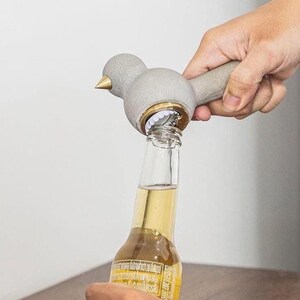 May include: A gray bird-shaped bottle opener with a brass beak and accents is opening a beer bottle. The opener is made of a solid material and has a smooth finish. The bottle is clear with a yellow liquid inside.