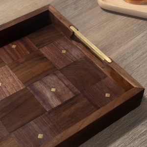 May include: A rectangular wooden serving tray with a geometric pattern of dark brown squares. The tray has a brass handle and small brass accents. The tray is designed for serving food and drinks.