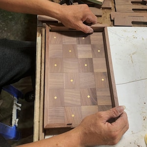 May include: A wooden chessboard with a checkerboard pattern of dark and light brown squares, each with a small gold-colored accent. The board is being assembled, with hands holding the frame.