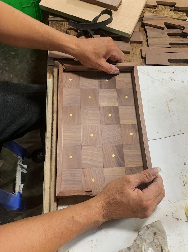 May include: A wooden chessboard with a checkerboard pattern of dark and light brown squares, each with a small gold-colored accent. The board is being assembled, with hands holding the frame.