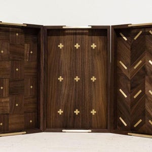 May include: A three-panel wooden tray set with a dark brown finish. The panels feature various inlaid patterns, including a patchwork design, crosses, and a chevron pattern. Gold-colored metal handles and accents are present.
