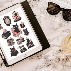 May include: An iPad displays a collection of gothic-themed stickers, including a black cat, a rose in a glass dome, and a bat. The stickers are in shades of black, pink, and gray. Sunglasses and a cocktail with a lemon slice are also visible.