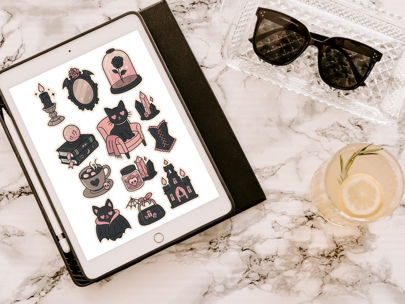 May include: An iPad displays a collection of gothic-themed stickers, including a black cat, a rose in a glass dome, and a bat. The stickers are in shades of black, pink, and gray. Sunglasses and a cocktail with a lemon slice are also visible.