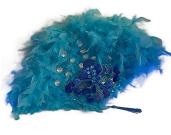Peut inclure: Éventail en plumes turquoise avec un motif décoratif. L'éventail présente un groupe de coquillages bleus et des ornements scintillants. Une gland pend du bas de l'éventail.