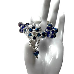 May include: A handcrafted bracelet featuring a floral design with blue and clear crystal beads. The bracelet has a silver chain and a dangling blue bead. The jewelry is displayed on a white hand.