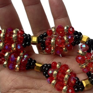 May include: A beaded bracelet with red, black, and gold beads. The bracelet has a spiral design with faceted red beads and gold-coloured accents. The clasp is gold-toned. The bracelet is shown against a white background.