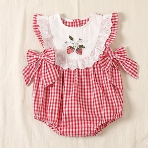 May include: Red and white gingham baby romper with a white bib featuring embroidered strawberries and eyelet lace trim. The romper has ruffled sleeves and bows at the shoulders, perfect for a summer outfit.