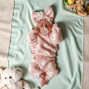 May include: A baby wearing a pink outfit with white floral patterns, including a sweater, pants, and a bow headband. The baby is also wearing a dummy. The baby is lying on a light green blanket with a teddy bear.