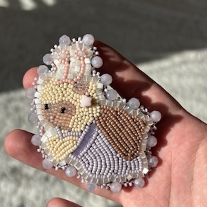 May include: A hand-beaded brooch featuring a whimsical bunny design. The brooch showcases a palette of soft colors, including beige, brown, lavender, and pink, with intricate beadwork. The bunny is adorned with a crown and surrounded by a beaded border.