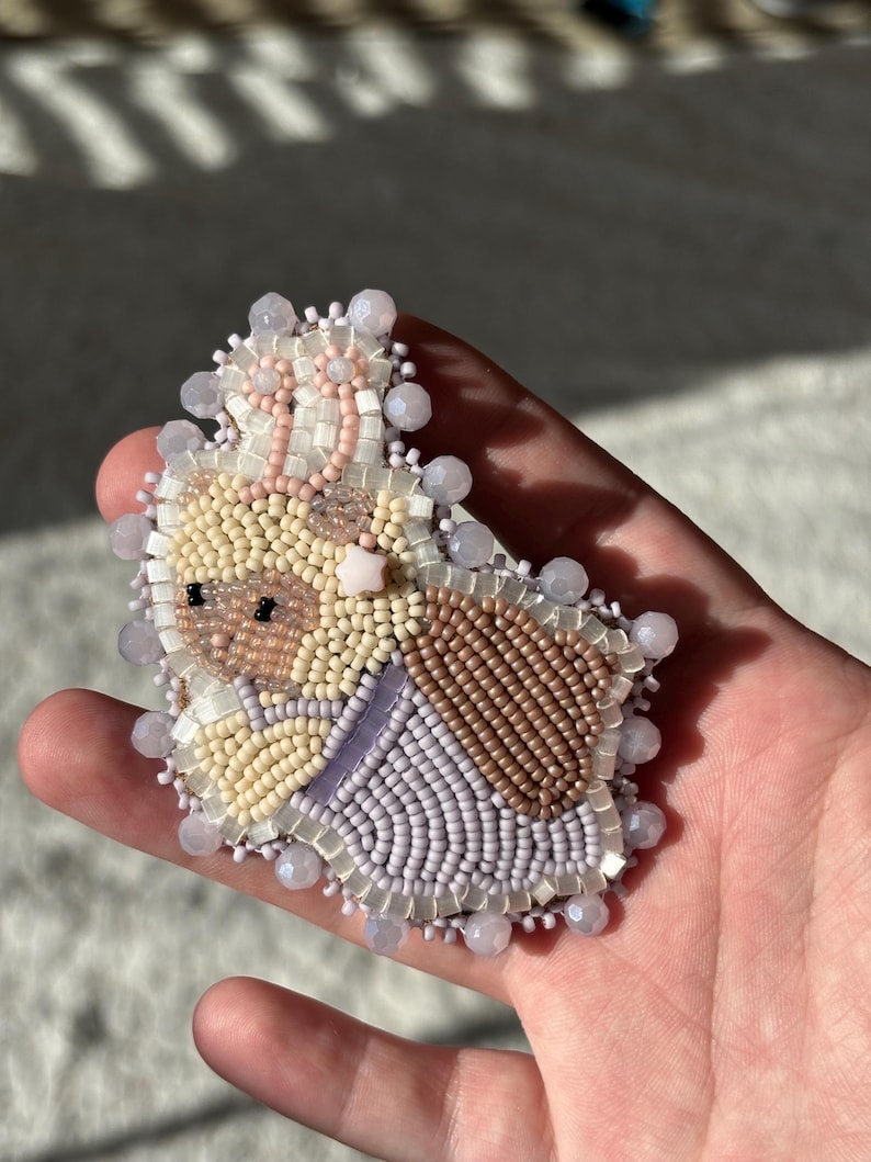 May include: A hand-beaded brooch featuring a whimsical bunny design. The brooch showcases a palette of soft colors, including beige, brown, lavender, and pink, with intricate beadwork. The bunny is adorned with a crown and surrounded by a beaded border.