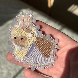 May include: A handmade beaded brooch in the shape of a bunny. The brooch features a variety of colors, including cream, tan, brown, lavender, and pink. The bunny has a detailed beaded face and is surrounded by a beaded border.