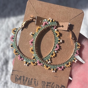 Op de afbeelding: Gouden oorringen met een kralen bloemenontwerp in pastelroze, geel en groen. De oorbellen zijn bevestigd aan een bruine kaart met de tekst "MUUU BEADS".