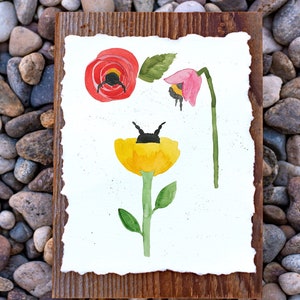 May include: A watercolor painting on paper mounted on a wooden frame. The painting features three flowers: a red poppy, a pink flower, and a yellow flower. Each flower has a black bumblebee on it.