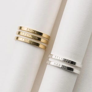 Peut inclure: Anneaux personnalisés en or et en argent avec des noms gravés. Les anneaux en or affichent les noms "EMMA", "GRAYSON" et "CHLOE". Les anneaux en argent affichent les noms "FELICITY" et "HARRIS". Les anneaux sont présentés sur un fond blanc.