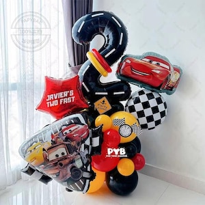 Puede incluir: Un arreglo festivo de globos con un gran globo negro con el número 2 diseñado como una pista de carreras. Incluye globos rojos, amarillos y negros, además de globos de personajes de la película Cars. Un globo estrella rojo dice "JAVIER'S TWO FAST".