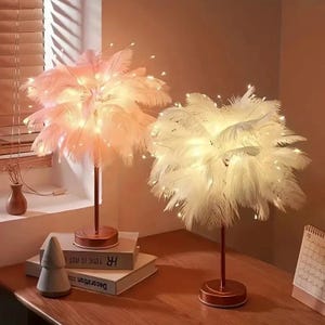May include: Two feather lamps with warm white LED lights. One lamp has a pink feather shade, the other has a white feather shade. Both lamps have a bronze-coloured base and stem. The lamps are on a wooden surface.