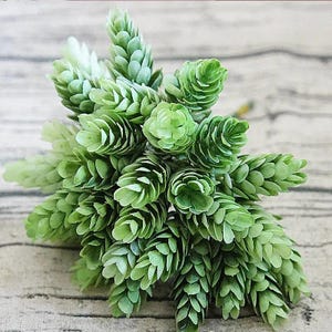 May include: A bunch of artificial green hops, with layered, cone-shaped blooms. The faux plant has varying shades of green, from light to deep, and is arranged against a light wood-grain background. This decorative item is suitable for home decor and craft projects.