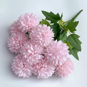 以下が含まれることがあります： 緑の葉と茎が付いた、薄いピンク色の人工的な菊の花の束。花は柔らかい花びらが何層にもなっており、ふっくらとした丸い形をしています。葉は濃い緑色です。