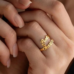 May include: A gold-colored ring with the names "Berta" and "Jessica" in a cursive font. The ring is wrapped around a finger, with the names displayed on the top of the finger. The ring is a personalized jewelry item.