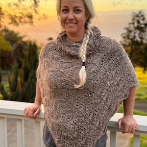 May include: A woman wearing a brown knitted poncho with a lace pattern. She has blonde hair braided down her back and is smiling at the camera.