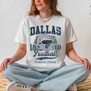 May include: White t-shirt with a distressed graphic featuring the word "DALLAS" above a football helmet and the text "1960 Football". Below the graphic is "DALLAS, TEXAS". The shirt is a casual style.