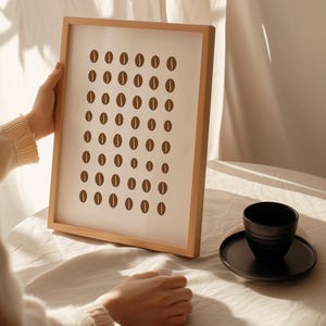 May include: A framed print with a pattern of brown coffee bean shapes on a white background, in a light wood frame. A black coffee cup and saucer are on a table. The image has a minimalist aesthetic.