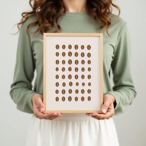 May include: A framed print featuring a minimalist design of coffee beans. The artwork showcases rows of brown coffee bean illustrations against a cream-colored background. The frame is a light wood color, adding a natural touch to the piece.