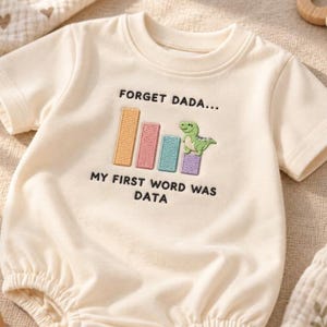 Könnte beinhalten: Ein cremefarbener kurzärmeliger Babybody mit einem gestickten Balkendiagramm und einem kleinen grünen Dinosaurier auf dem letzten Balken. Der Text FORGET DADA... MY FIRST WORD WAS DATA ist in schwarzer Stickerei über und unter dem Motiv.