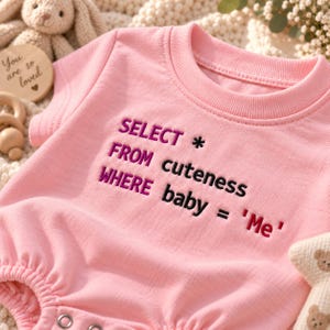 Puede incluir: Un body para bebé de color rosa claro con el texto bordado: SELECT * FROM cuteness WHERE baby = 'Me'. La prenda tiene un cuello redondo acanalado y mangas cortas, con broches de presión metálicos visibles en el dobladillo inferior.