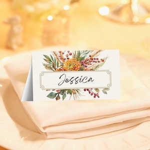 May include: A place card with the name "Jessica" in elegant script, framed by a decorative border. The card features a watercolor floral design with orange, green, and red accents. It rests on a folded beige napkin atop a white plate.