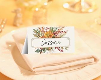 May include: A place card with the name "Jessica" in elegant script, framed by a decorative border. The card features a watercolor floral design with orange, green, and red accents. It rests on a folded beige napkin atop a white plate.