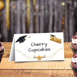 May include: A graduation-themed display featuring cherry cupcakes and a sign that reads "Cherry Cupcakes" with a graduation cap and the year 2026. The cupcakes have pink frosting, chocolate shavings, and a cherry on top. The sign is on a wooden surface.