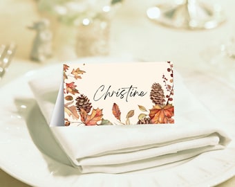 May include: A cream-coloured place card featuring the name "Christine" in script. It's adorned with watercolour illustrations of autumn leaves, pine cones, and berries, ideal for a seasonal celebration.