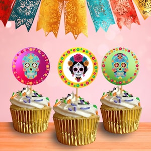 May include: Three cupcakes with white frosting and colorful sprinkles, topped with decorative sugar skull toppers. The toppers are round with floral and skull designs in pink, green, and yellow. Gold cupcake liners.