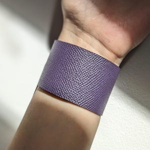 Può includere: Un bracciale a polsino largo in pelle viola strutturata. Il bracciale è indossato al polso e ha una superficie martellata. Il bracciale è di colore pieno e sembra essere realizzato in pelle di alta qualità. Il bracciale è un accessorio audace.